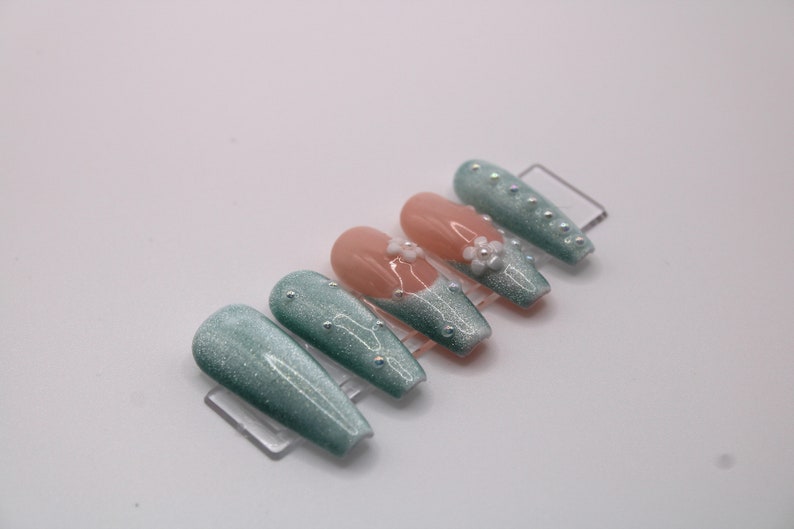 May include: Set of ten press-on nails with a teal glitter finish. Some nails have a white flower accent with pearl embellishments.