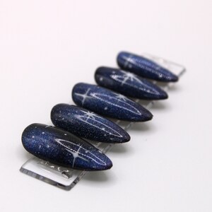 May include: A set of six blue and white nail tips with a galaxy design. The tips are arranged in a row on a clear plastic display stand.