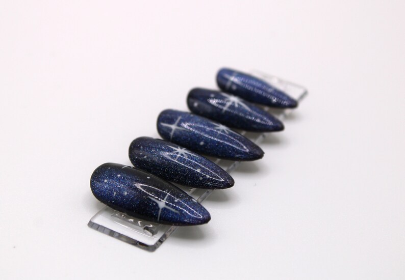 May include: A set of six blue and white nail tips with a galaxy design. The tips are arranged in a row on a clear plastic display stand.