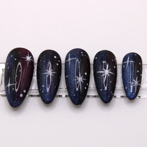 May include: Set of six false nails with a dark blue and purple galaxy design with white glitter and stars.