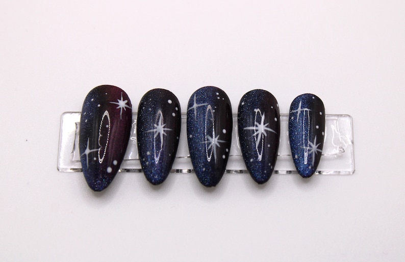 May include: Set of six false nails with a dark blue and purple galaxy design with white glitter and stars.