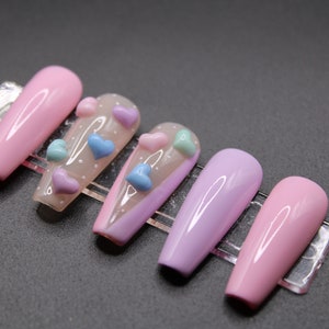 Op de afbeelding: Set van zes lange, roze en paarse kunstnagels met een transparante basis en 3D-hartversieringen in roze, blauw en lichtblauw.