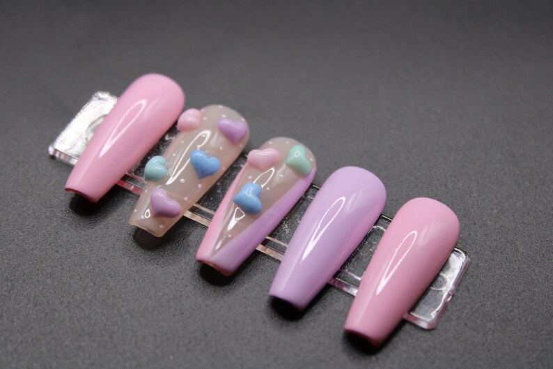 Op de afbeelding: Set van zes lange, roze en paarse kunstnagels met een transparante basis en 3D-hartversieringen in roze, blauw en lichtblauw.