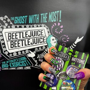 May include: A black and green Beetlejuice themed button set with the text "BUTTONS". The background features the text "THE GHOST WITH THE MOST!" and "BEETLEJUICE". The image also shows a hand with nail art.