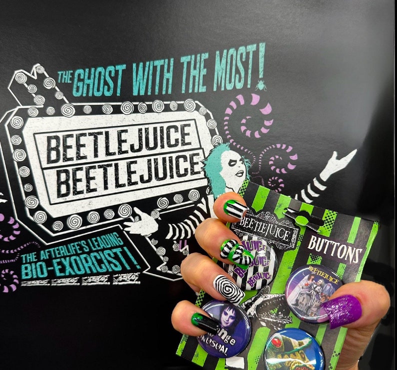May include: A black and green Beetlejuice themed button set with the text "BUTTONS". The background features the text "THE GHOST WITH THE MOST!" and "BEETLEJUICE". The image also shows a hand with nail art.
