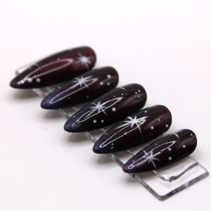 May include: Set of six long, dark purple and black stiletto false nails with white star and dot designs.