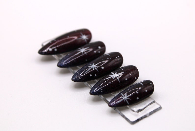 May include: Set of six long, dark purple and black stiletto false nails with white star and dot designs.