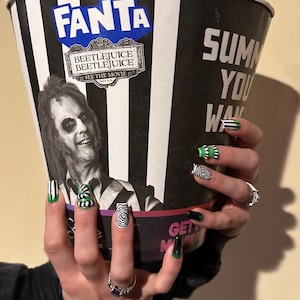 May include: A large black and white striped popcorn bucket with the Fanta and Beetlejuice logos. The bucket is held up by a person with black, green, and white nail art. The nails feature stripes, swirls, and the words "Get Movie".