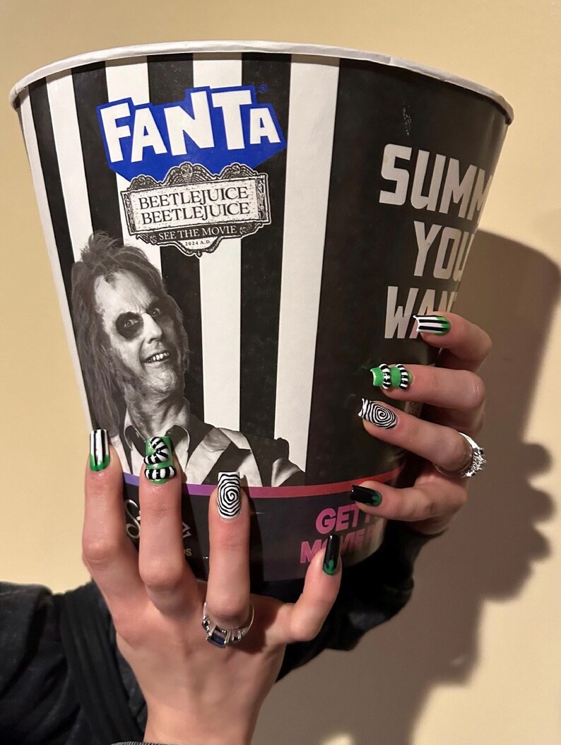 May include: A large black and white striped popcorn bucket with the Fanta and Beetlejuice logos. The bucket is held up by a person with black, green, and white nail art. The nails feature stripes, swirls, and the words "Get Movie".