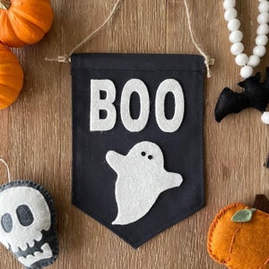 May include: A black felt banner with the word "BOO" in white felt letters and a white felt ghost with two black eyes. The banner is hanging from a string and is surrounded by Halloween decorations.