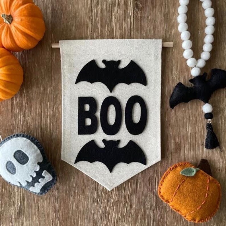 May include: A white felt banner with the word "BOO" in black felt letters and two black felt bats. The banner is hanging from a wooden dowel rod.