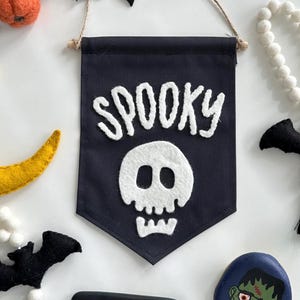 May include: A black felt banner with white felt lettering that reads "SPOOKY" and a white felt skull. The banner is hanging from a string and has a wooden dowel at the top.