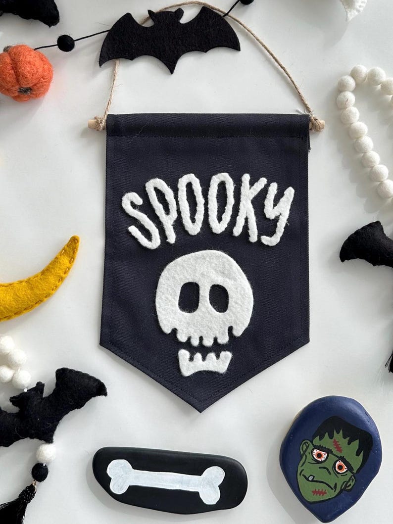 May include: A black felt banner with white felt lettering that reads "SPOOKY" and a white felt skull. The banner is hanging from a string and has a wooden dowel at the top.