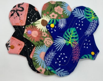 May include: Four reusable cloth menstrual pads with various patterns. One pad features a black background with pink accents and a celestial design. Another has a floral pattern in shades of green and pink. The third pad has a blue background with white leaves. Each pad has snap closures.
