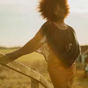 May include: A woman with curly brown hair wearing a grey knitted sweater and a brown skirt stands in a field, leaning on a wooden fence.