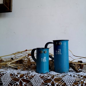 Könnte beinhalten: Zwei blaue Emaille-Messbecher mit Griffen. Der kleinere Becher ist mit "1 dL" und der größere mit "2 dL" beschriftet. Die Becher stehen auf einer weißen Häkeltischdecke.