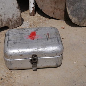 Op de afbeelding: Een rechthoekige, zilverkleurige metalen EHBO-kit met een rood kruis op de bovenkant. De kit heeft een scharnierend deksel en een sluiting met een sluiting. De tekst op de bovenkant van de kit is gedeeltelijk zichtbaar.