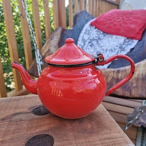 May include: A bright red enamel teapot with a black lid and handle. The teapot is sitting on a wooden surface.