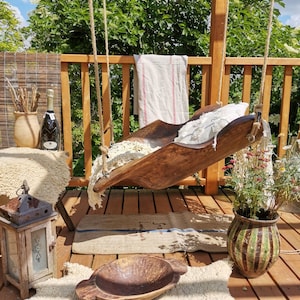 May include: A wooden swing seat hangs from a wooden pergola. The swing seat is made of a wooden trough with a white fabric cushion. The swing seat is hanging over a wooden deck with a rug and a wooden bowl.