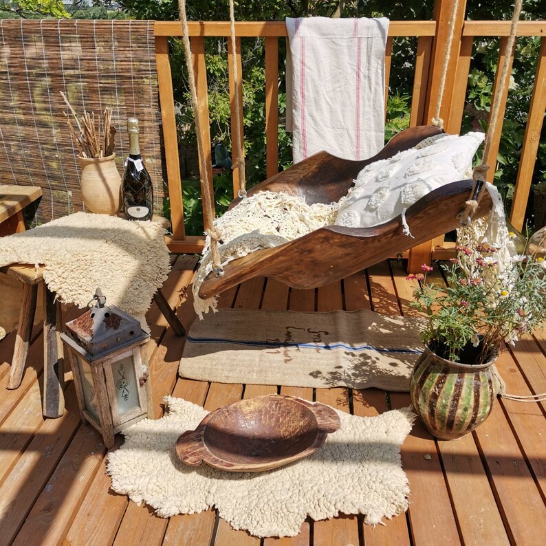 May include: A wooden swing seat with a white crocheted cushion and a white and red striped fabric hanging behind it. The swing is hanging from a wooden frame and is surrounded by plants and a wooden floor.