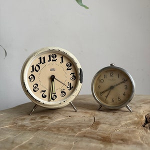 May include: Two vintage alarm clocks, one with a white face and black numbers, the other with a silver face and black numbers. Both clocks have black hands and are on a wooden surface.