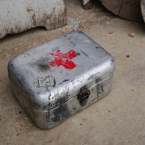 May include: A silver metal first aid kit with a red cross on the lid. The kit has a worn, vintage look and is slightly dented.
