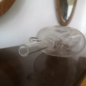 May include: A clear glass bottle with a long, thin spout and a rounded body. The bottle is lying on its side on a dark brown surface.