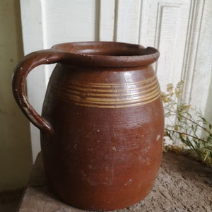 Può includere: Un'anfora di terracotta marrone con un'ampia apertura e un manico. L'anfora ha un aspetto rustico e fatto a mano ed è decorata con strisce marroni.