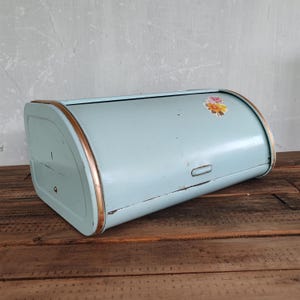 Puede incluir: Panera vintage de metal azul claro con tapa enrollable y ribete color cobre. Una pegatina floral está en el lateral. La panera está sobre una superficie de madera.