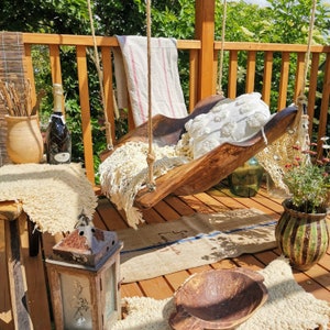 May include: A wooden swing seat with a white crocheted cushion and a white crocheted throw blanket. The swing is hanging from a wooden pergola with a wooden railing. There is a bottle of wine and a lantern on the ground.