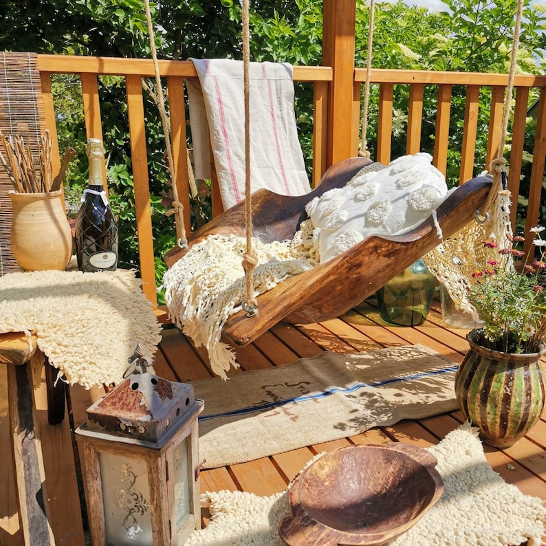 May include: A wooden swing seat with a white crocheted cushion and a white crocheted throw blanket. The swing is hanging from a wooden pergola with a wooden railing. There is a bottle of wine and a lantern on the ground.