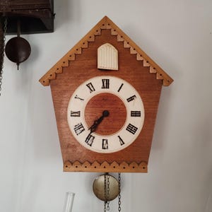 May include: A wooden cuckoo clock shaped like a house with a white face and black Roman numeral hour markers. The clock has a pendulum and a small weight hanging from chains.