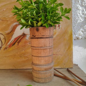 Könnte beinhalten: Ein rustikale Holzvase, etwa 20 cm hoch, gefüllt mit frischen grünen Zweigen. Die Vase hat eine natürliche Holzoberfläche mit horizontalen Bändern. Der Hintergrund zeigt ein Gemälde in warmen Farben, was eine natürliche Ästhetik erzeugt.