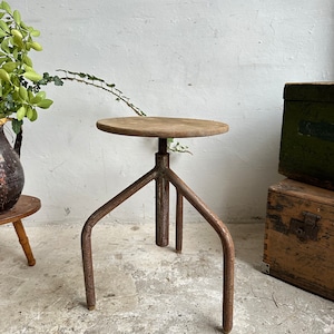 Puede incluir: Un taburete de madera rústico con una tapa redonda y tres patas de metal. El taburete es de altura ajustable y tiene un aspecto desgastado y vintage.