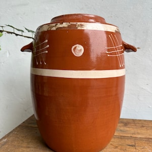 Puede incluir: Una jarra de terracota con diseños pintados en blanco. La jarra tiene dos pequeñas asas a los lados y una banda blanca alrededor de la parte superior.