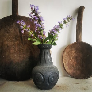 May include: A black, textured vase with a fluted neck and scrollwork design, holding purple flowers with green leaves. Two large, wooden, antique kitchen utensils flank the vase, creating a rustic, farmhouse aesthetic.