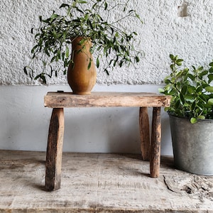 May include: A rustic, wooden bench with four sturdy legs. A brown, antique vase filled with green foliage sits on the bench. A metal bucket with green plants is to the right. The bench is made of weathered wood.