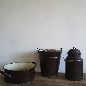 Puede incluir: Tres artículos de cocina antiguos de esmalte marrón sobre una superficie de madera. Incluye una olla ovalada grande con asas, un cubo con asa de madera y una lechera con tapa y asa de madera. Los artículos están contra una pared blanca.
