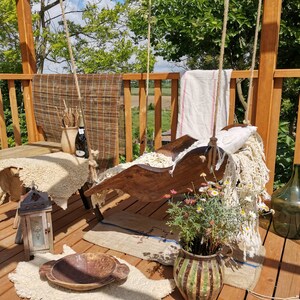 May include: A wooden swing seat with white fabric cushions and a white throw blanket. The swing is hanging from a wooden pergola with a view of a green field and trees in the background. There are flowers in a pot in the foreground.