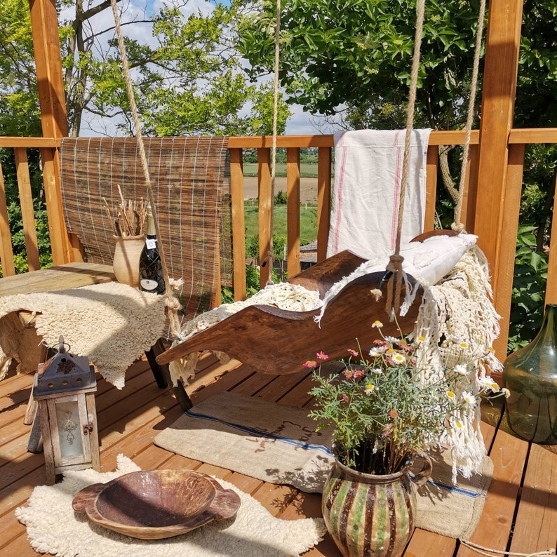 May include: A wooden swing seat with white fabric cushions and a white throw blanket. The swing is hanging from a wooden pergola with a view of a green field and trees in the background. There are flowers in a pot in the foreground.