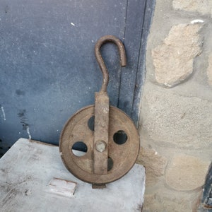 May include: A rusty metal pulley with a hook, featuring four holes in the wheel. The pulley is on a light gray surface.