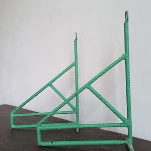 May include: Two green metal shelf brackets with a geometric design. The brackets are made of rebar and have a rustic, industrial look.