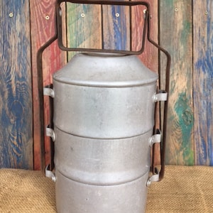 Op de afbeelding: Een vintage, zilveren, driedelige lunchbox met een metalen handvat. De lunchbox staat op een bruine jute ondergrond.