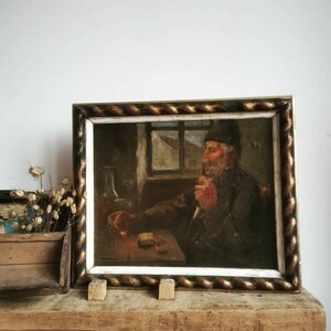 May include: A framed oil painting of a man with a beard sitting in a room, holding a pipe in his hand. The painting is in a gold and white frame with a rope-like design.