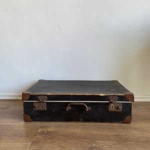 Pode incluir: Uma mala vintage preta com detalhes em couro marrom e fechos de metal. A mala tem uma alça na parte superior e está sobre um piso de madeira.