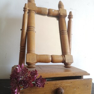 May include: A wooden vanity mirror with a small drawer. The mirror is attached to a stand with a decorative design. The mirror is surrounded by a wooden frame. The drawer is made of wood and has a small knob. The mirror is sitting on a dark surface.