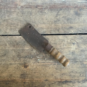 May include: A rusty cleaver with a wooden handle. The cleaver is lying on a wooden surface.