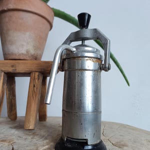 May include: A vintage silver and black stovetop espresso maker with a lever handle. The maker has a worn, aged look with some rust spots.