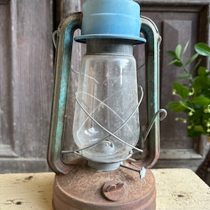 Op de afbeelding: Een vintage, roestige, groene en blauwe olielamp met een glazen schoorsteen en een metalen kooi. De lamp heeft een blauwe metalen bovenkant en een roestige metalen basis.