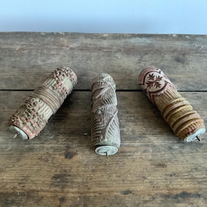 May include: Three vintage wooden paint rollers with intricate patterns. The rollers are brown, beige, and red with a white background. Each roller has a metal pin at the end.